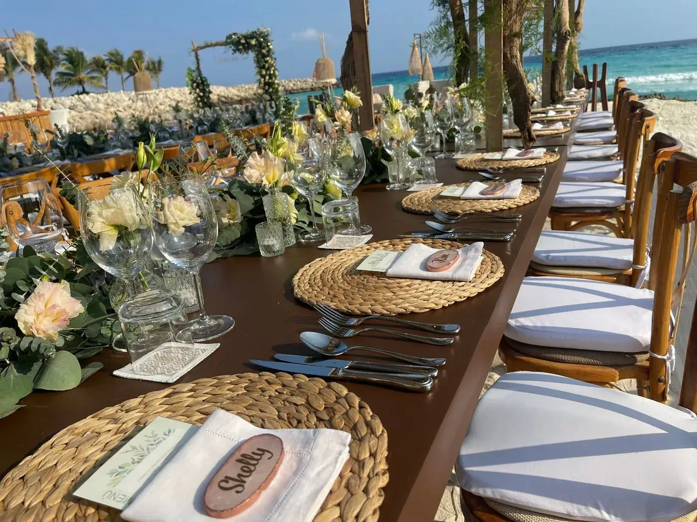 Beachfront destination wedding reception table with rattan placemats, eucalyptus runner, and ocean view
