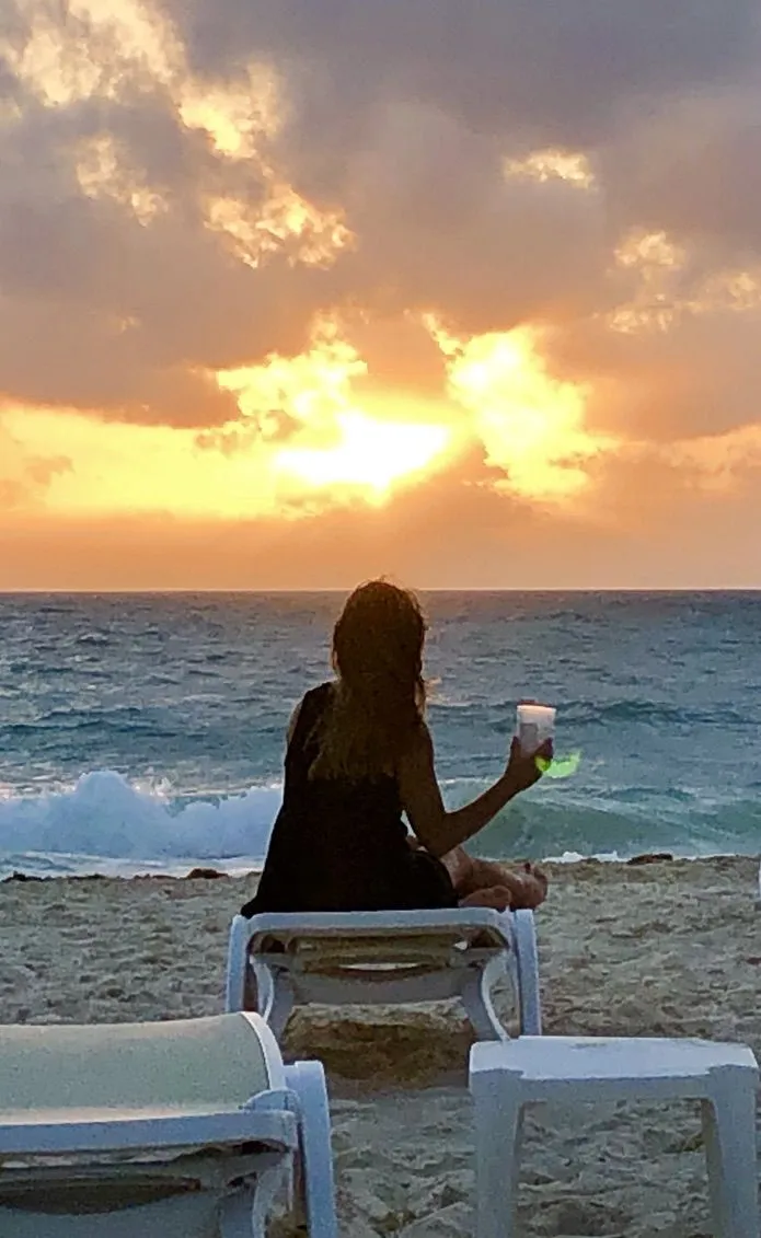 Woman watching a golden Caribbean sunset from a beach lounger with a cocktail in hand