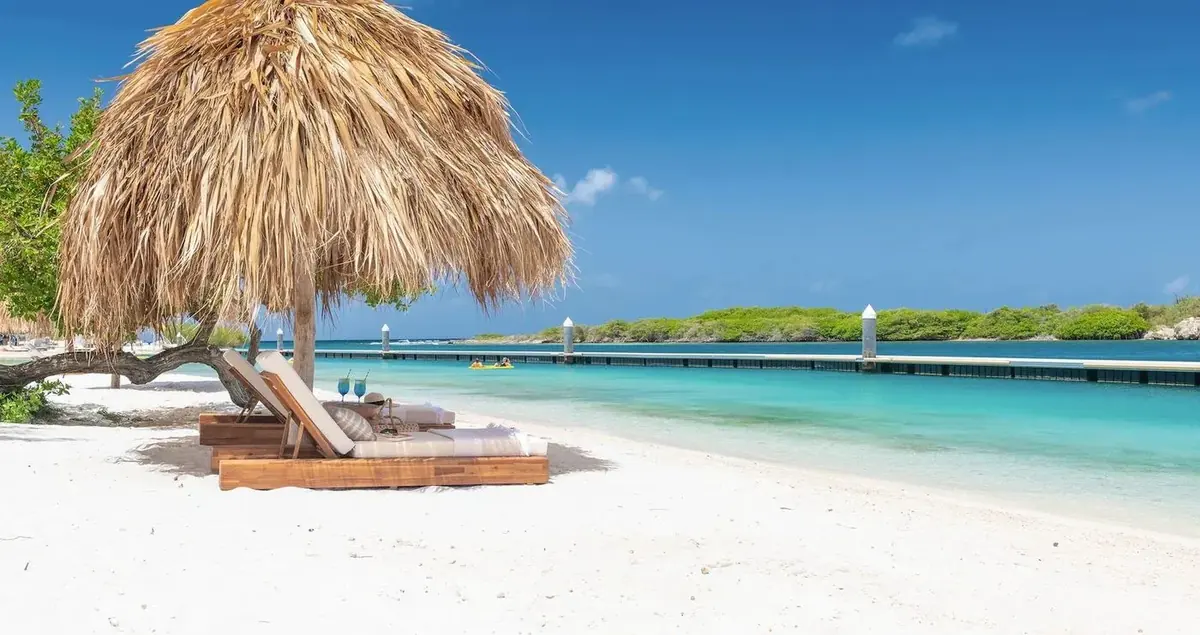 Beachside lounge chairs under a palapa umbrella at Sandals Royal Curaçao with crystal clear turquoise water